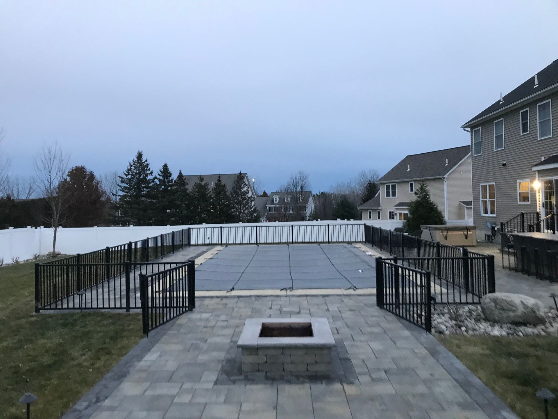 Backyard with pool covered, fire pit, patio, fence, and houses in the distance. Overcast sky.