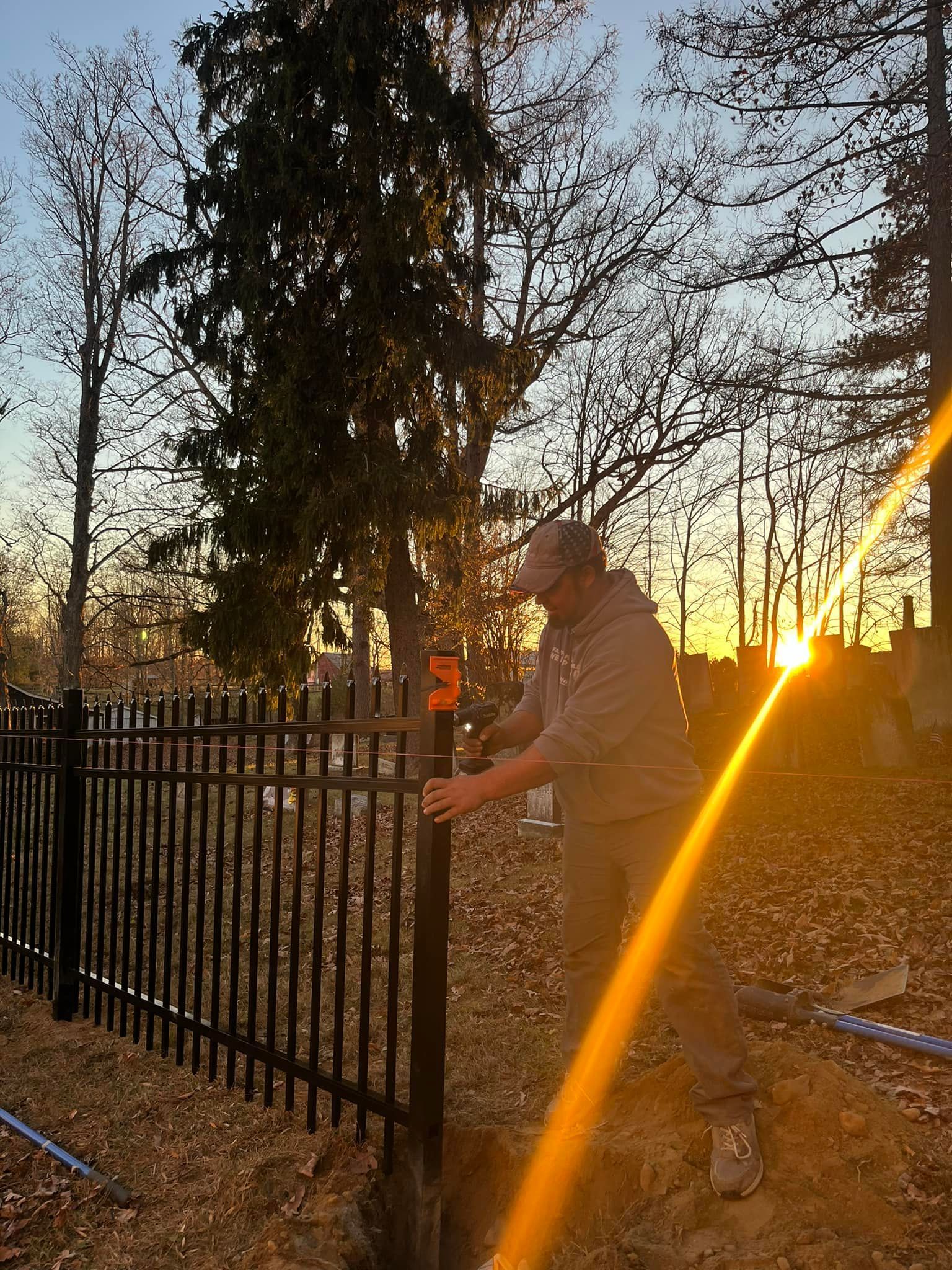 Man installing black metal fence post in a trench, sun setting in background.
