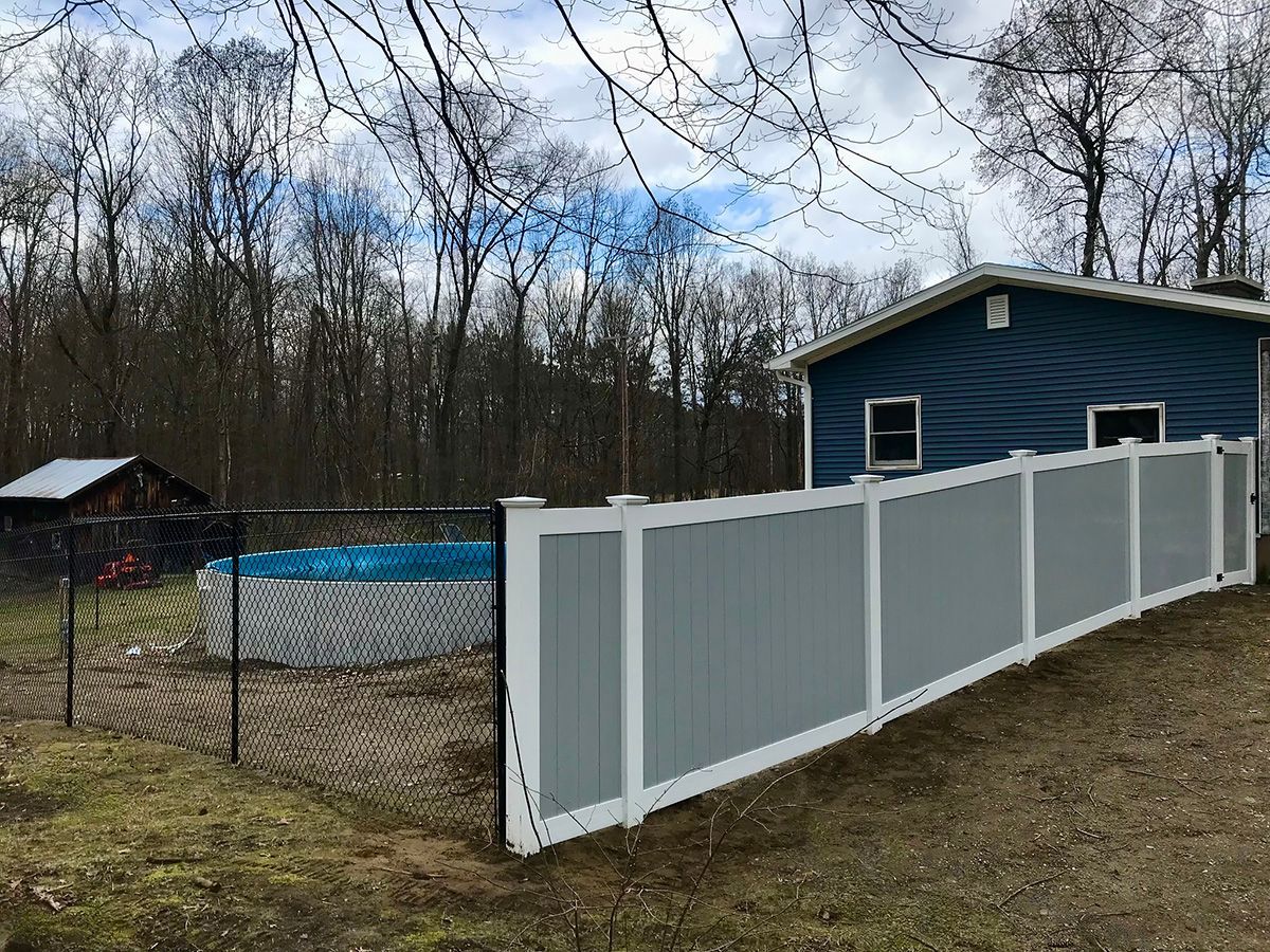 A gray and white privacy fence surrounds a blue-sided house and pool, with trees in the background.