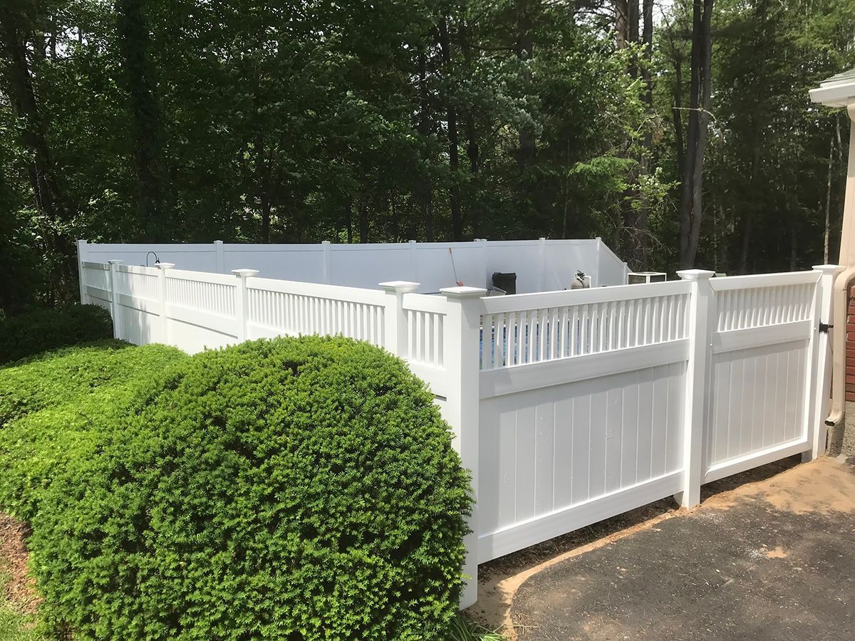 White vinyl fence encloses a backyard with trees and a large green bush.
