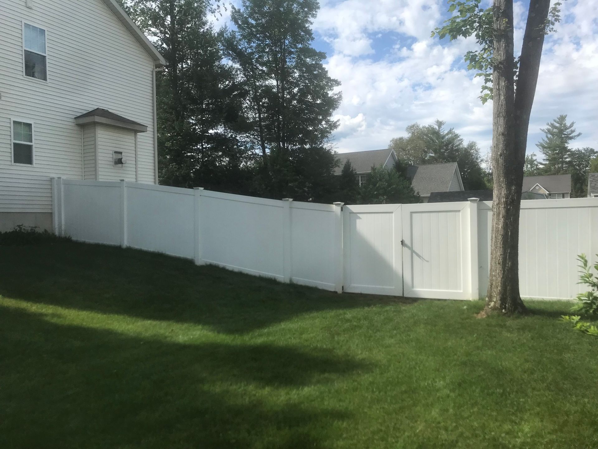 White vinyl fence with gate in a backyard, beside a house and trees, on a sunny day.