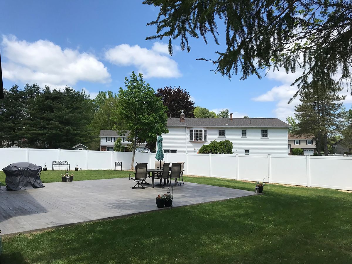 Backyard with patio, lawn, white fence, and house under a blue sky with clouds.