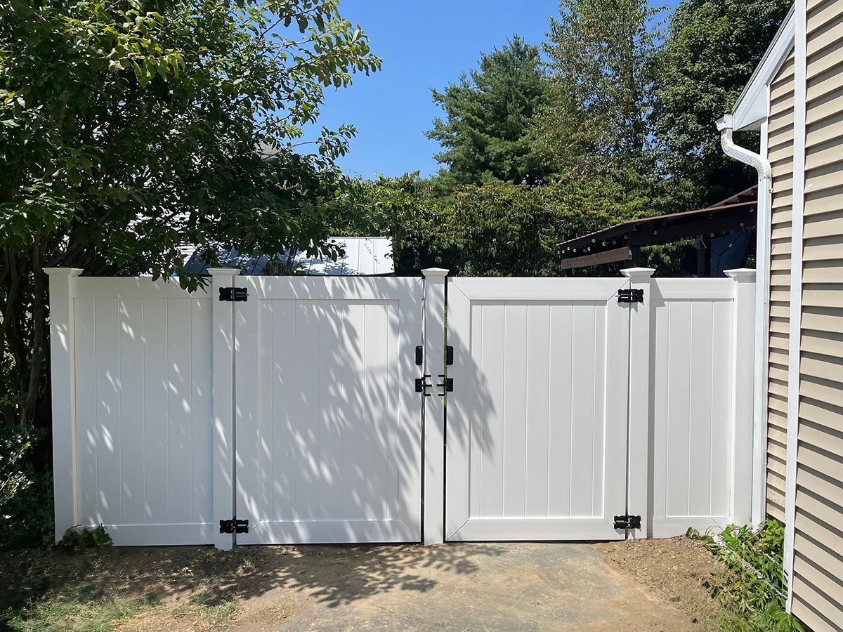 White vinyl fence with double gates, black hardware, set against trees and a building on a sunny day.