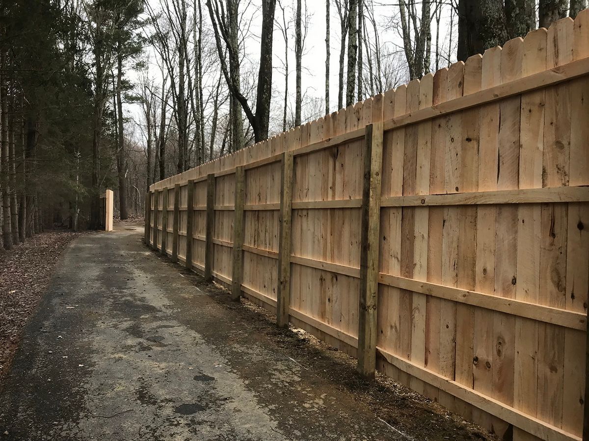 A wooden fence lines a gravel path through a wooded area. The fence is light brown.