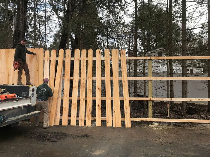Two people constructing a wooden fence outdoors, one on a truck bed, in front of a wooded backdrop.