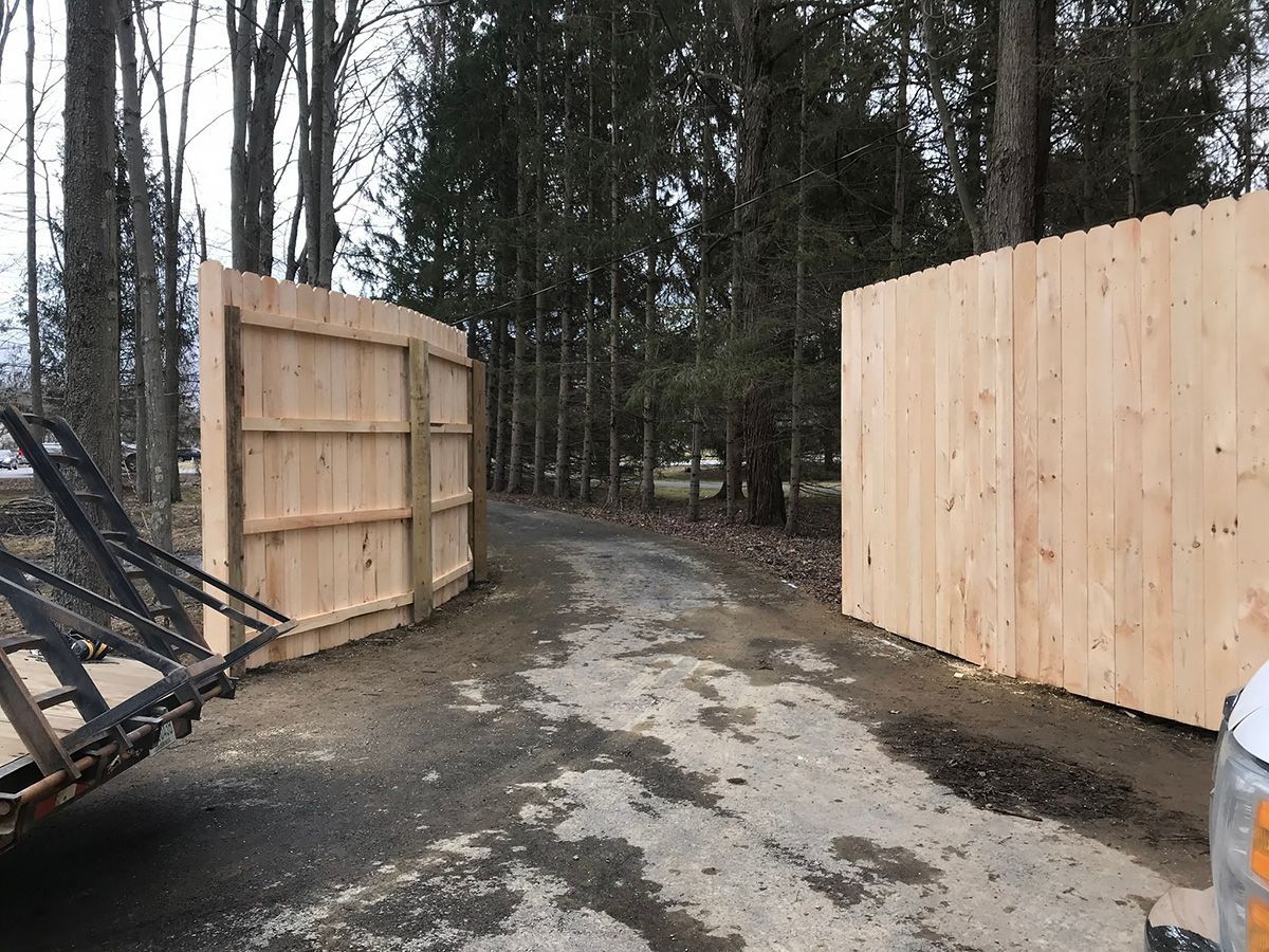 Open wooden gate, gravel driveway, and unfinished fence in a wooded area.