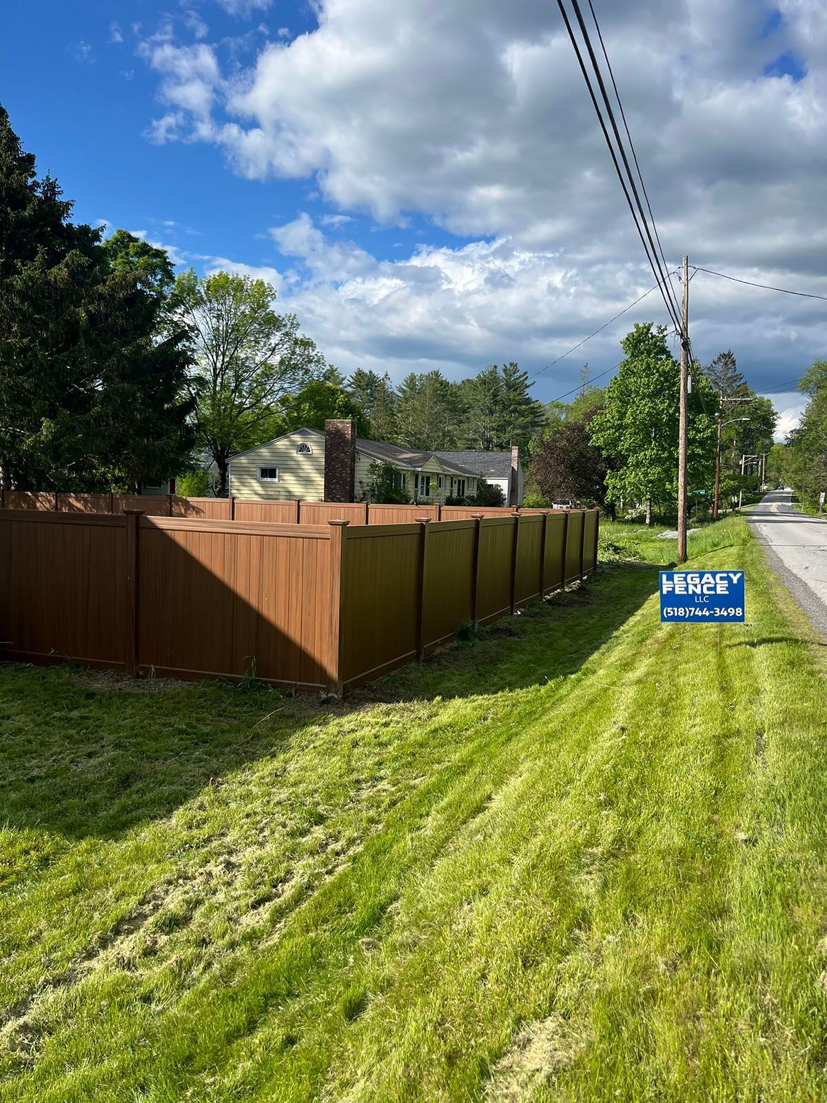Brown privacy fence alongside a grassy roadside, with a house visible in the background under a blue sky.