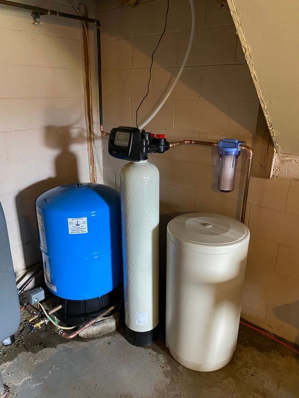Water treatment system in a basement. Includes a blue tank, tall white tank, and a white salt tank.