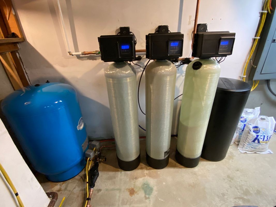 Three water filtration systems with digital controls, next to a blue water tank in a basement.