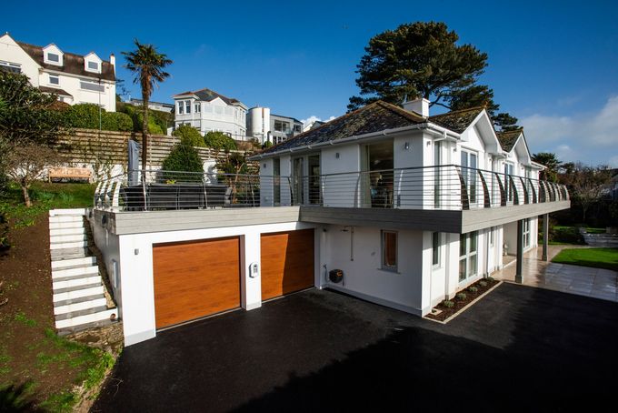 A modern white house with a two-car garage, a raised deck with metal railings, and a paved driveway on a sunny day.