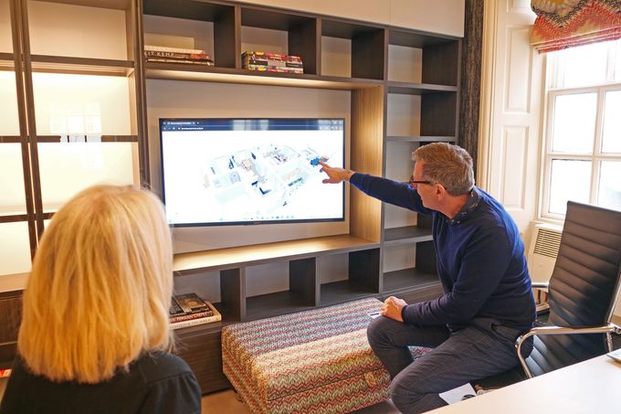 A professional sitting in an office points at a complex diagram displayed on a wall-mounted television screen.