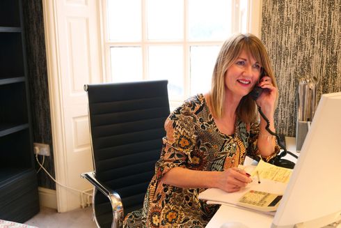 A person with shoulder-length hair wearing a patterned dress sits at a desk on the phone while writing in a notebook.