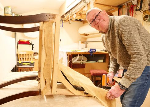 A person in a workshop uses a staple gun to attach fabric to the wooden frame of a chair undergoing reupholstery.