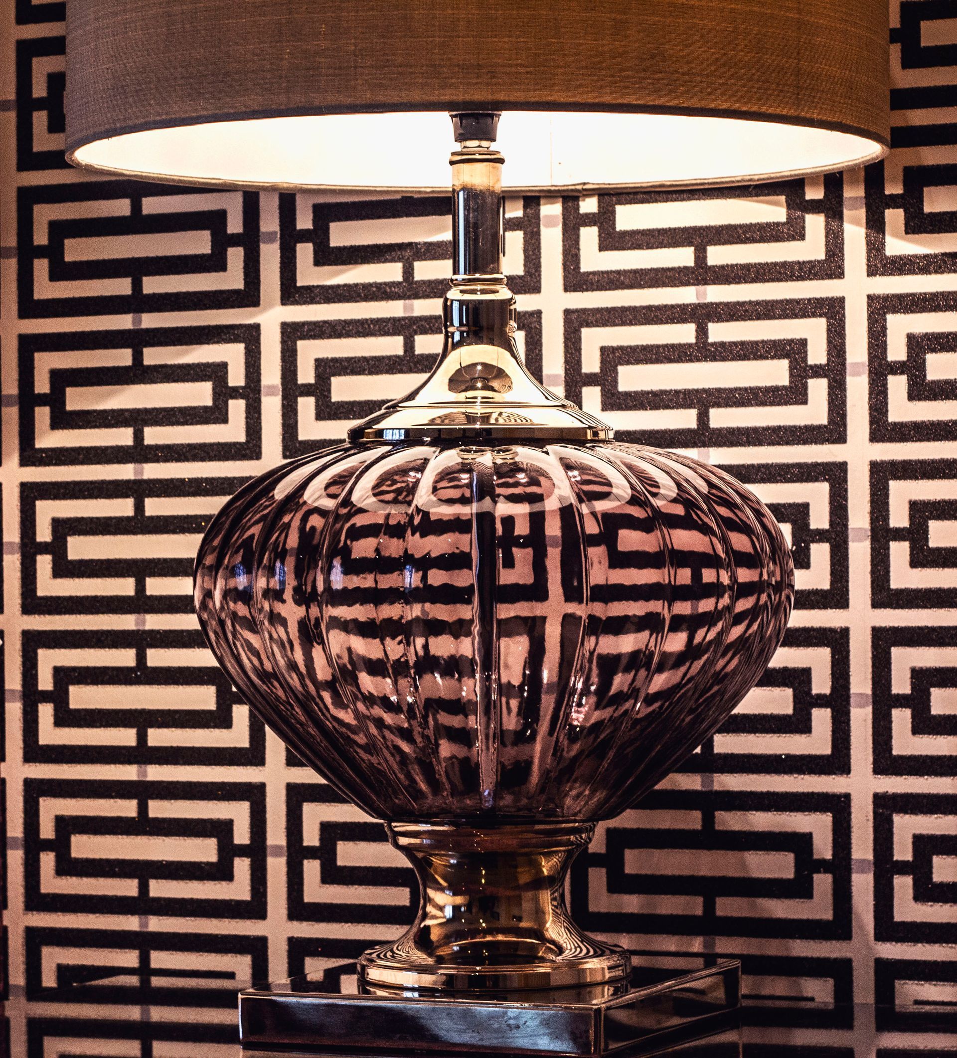 A table lamp with a dark, ridged glass base and a matching shade set against a black-and-white geometric patterned wall.
