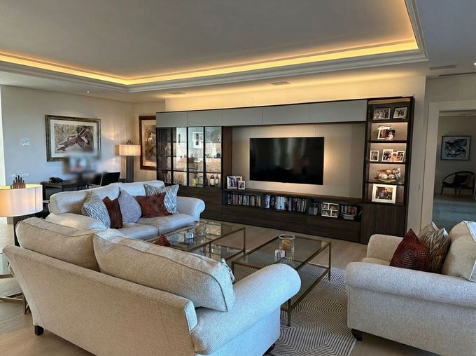 A contemporary living room featuring a large beige sofa, a matching armchair, a central TV wall unit, and warm lighting.