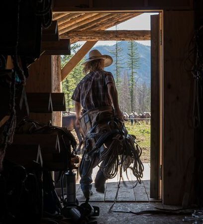 A man in a cowboy hat is walking out of a barn with a horse.