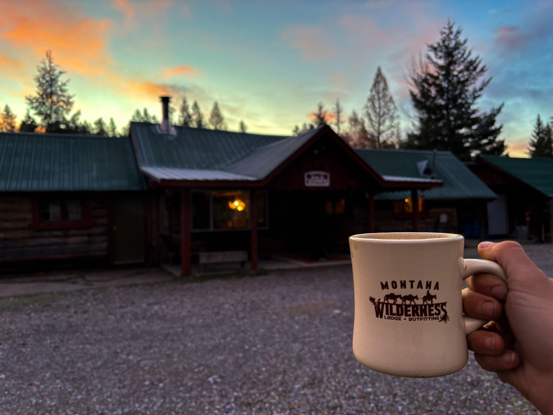 A person is holding a montana wilderness coffee mug in front of a cabin.