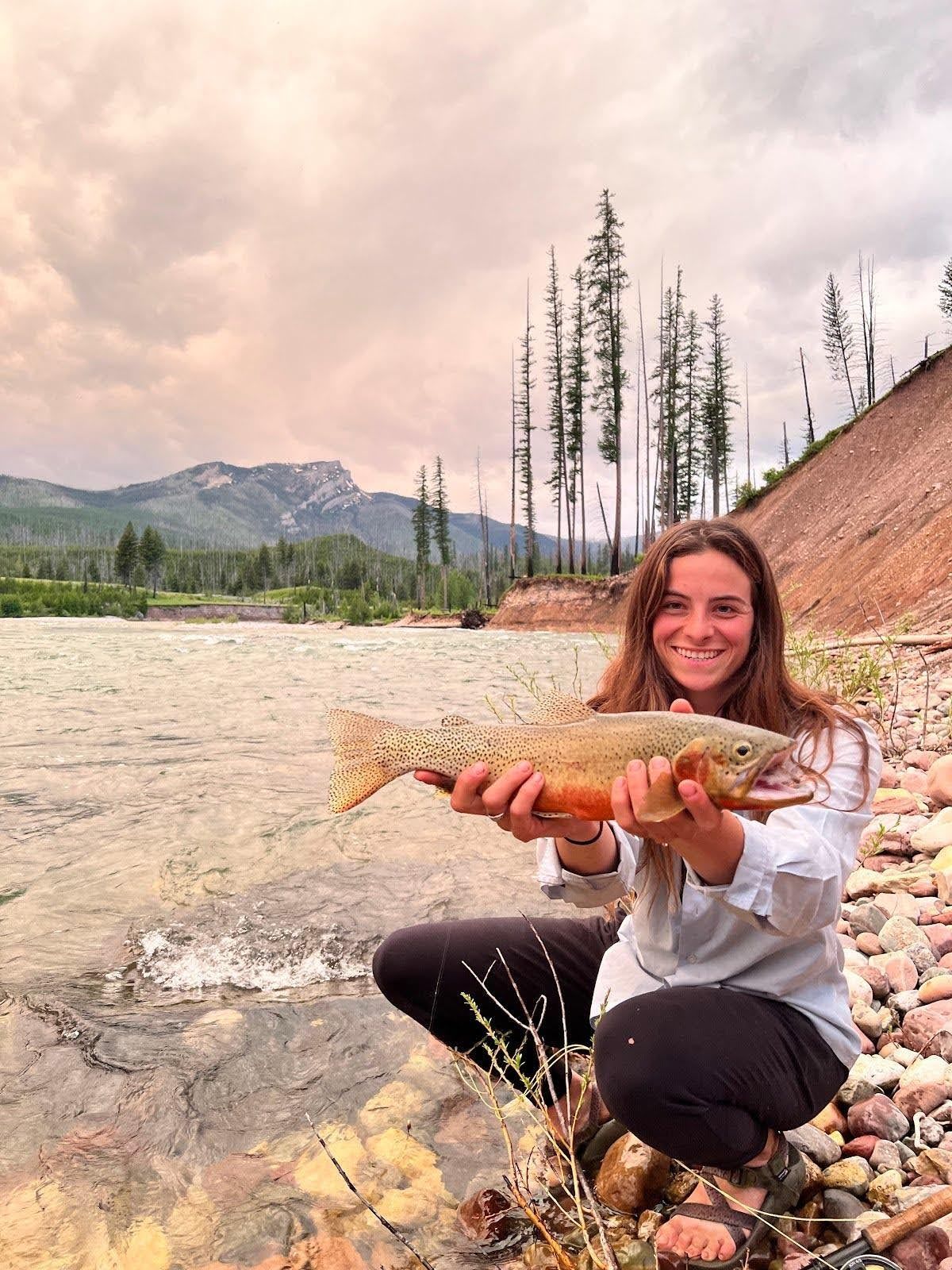 A woman is kneeling down next to a river holding a fish.