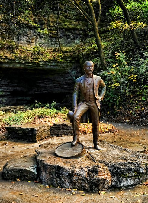 Bronze statue of a man in a suit standing on a rock with a barrel, set in a wooded area.