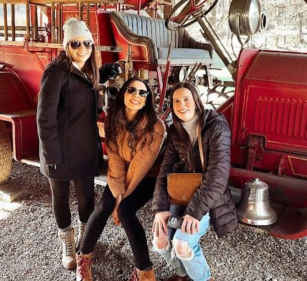Three women smiling next to a vintage red fire truck. They are wearing winter coats, and one is sitting on the truck.