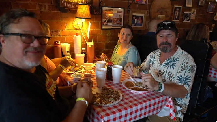 A man in a black polo shirt and sunglasses sings into a microphone. An acoustic guitar and a 