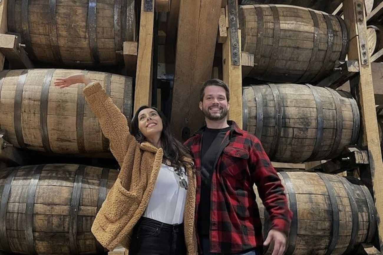 A woman with one arm raised and a man stand in front of a stack of wooden barrels. They smile in a dimly lit room.