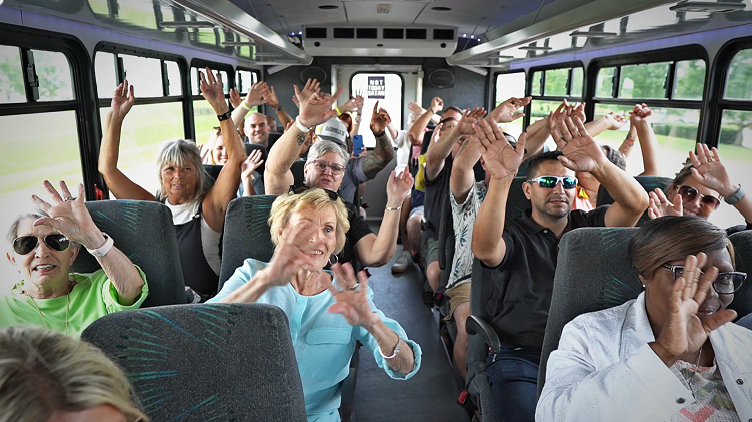 A group of people are sitting on a bus, smiling with their hands in the air.