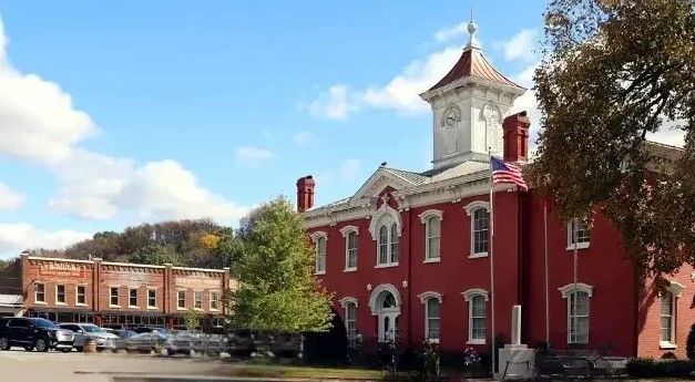 Red brick courthouse with clock tower and American flag on a sunny day. Trees and parked cars are in front.