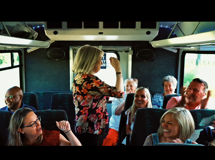 Woman leading a group on a bus, raising her fist, dancing, people smiling and laughing.