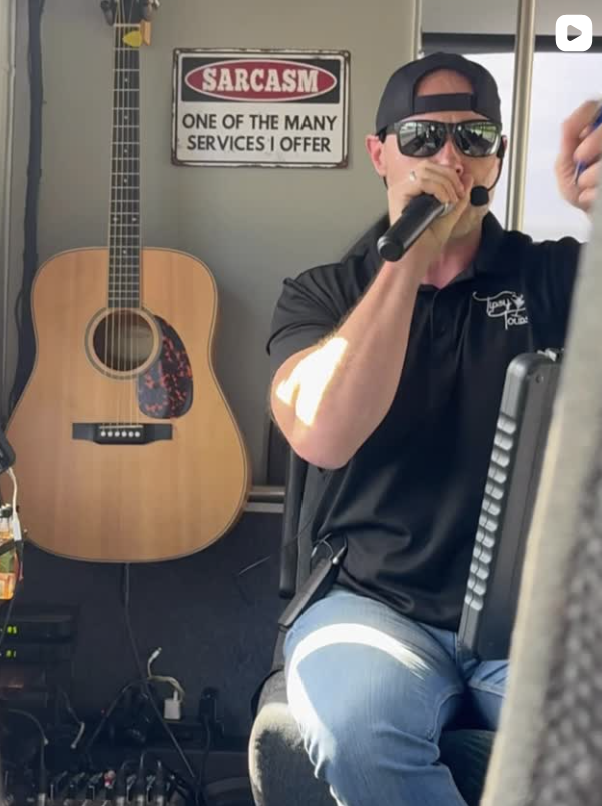Man in sunglasses sings into a microphone on a bus, guitar beside him. A 