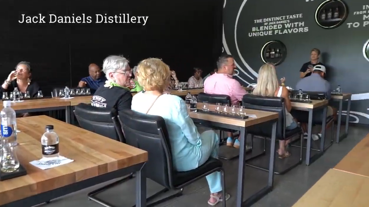 A group of people at the Jack Daniel's Distillery tasting session, listening to a presenter. Tables hold glasses of whiskey.