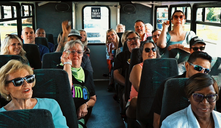 People of various ages on a bus. Several are wearing sunglasses, looking toward the camera.