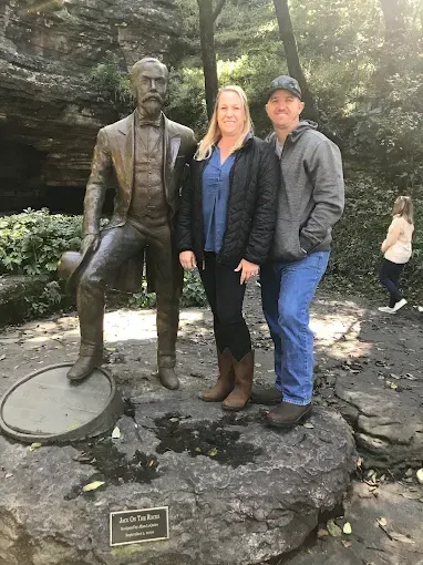 Couple posing with a statue of a man in front of a rock wall and greenery. The woman wears a blue shirt, black jacket.