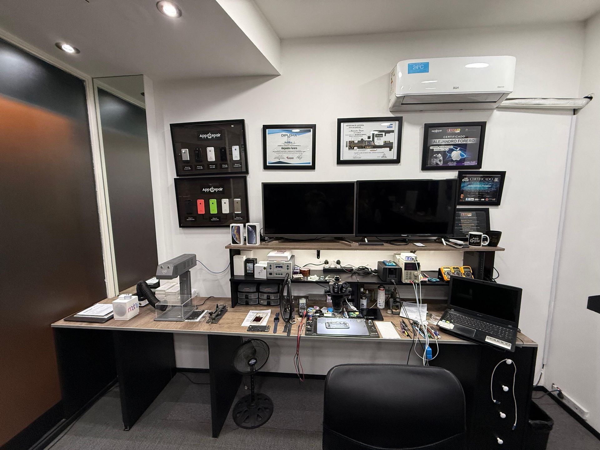 Un espacio de trabajo de taller de reparaciones con aparatos electrónicos, herramientas y certificados enmarcados.