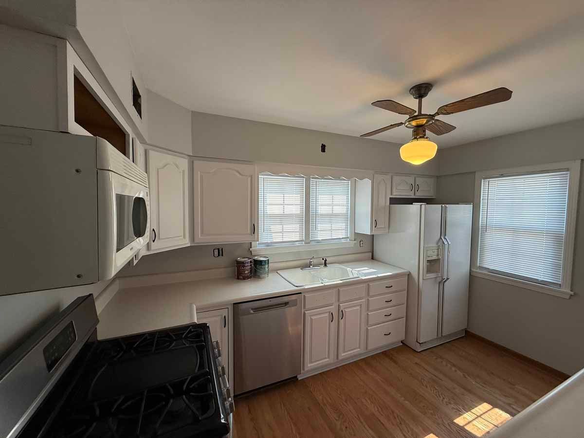 White kitchen with appliances, cabinets, and a ceiling fan. Wooden floors and windows provide natural light.
