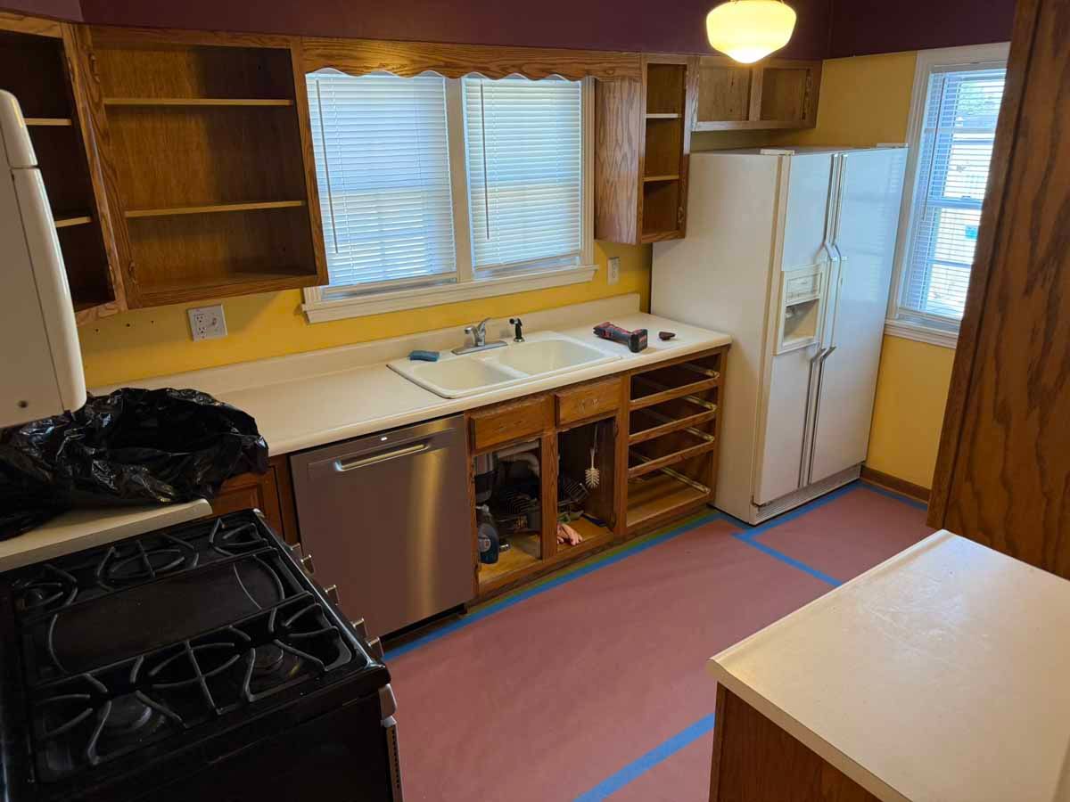 Kitchen undergoing renovation; yellow walls, wooden cabinets, appliances and white countertops.