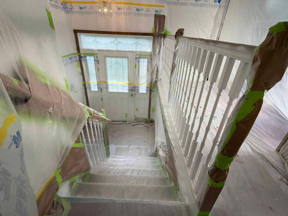 Staircase during renovations, covered in plastic sheeting and painter's tape. White railing, doorway at the top.