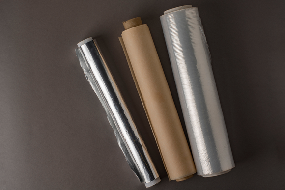 Three rolls of aluminum foil , plastic wrap , and brown paper on a table.