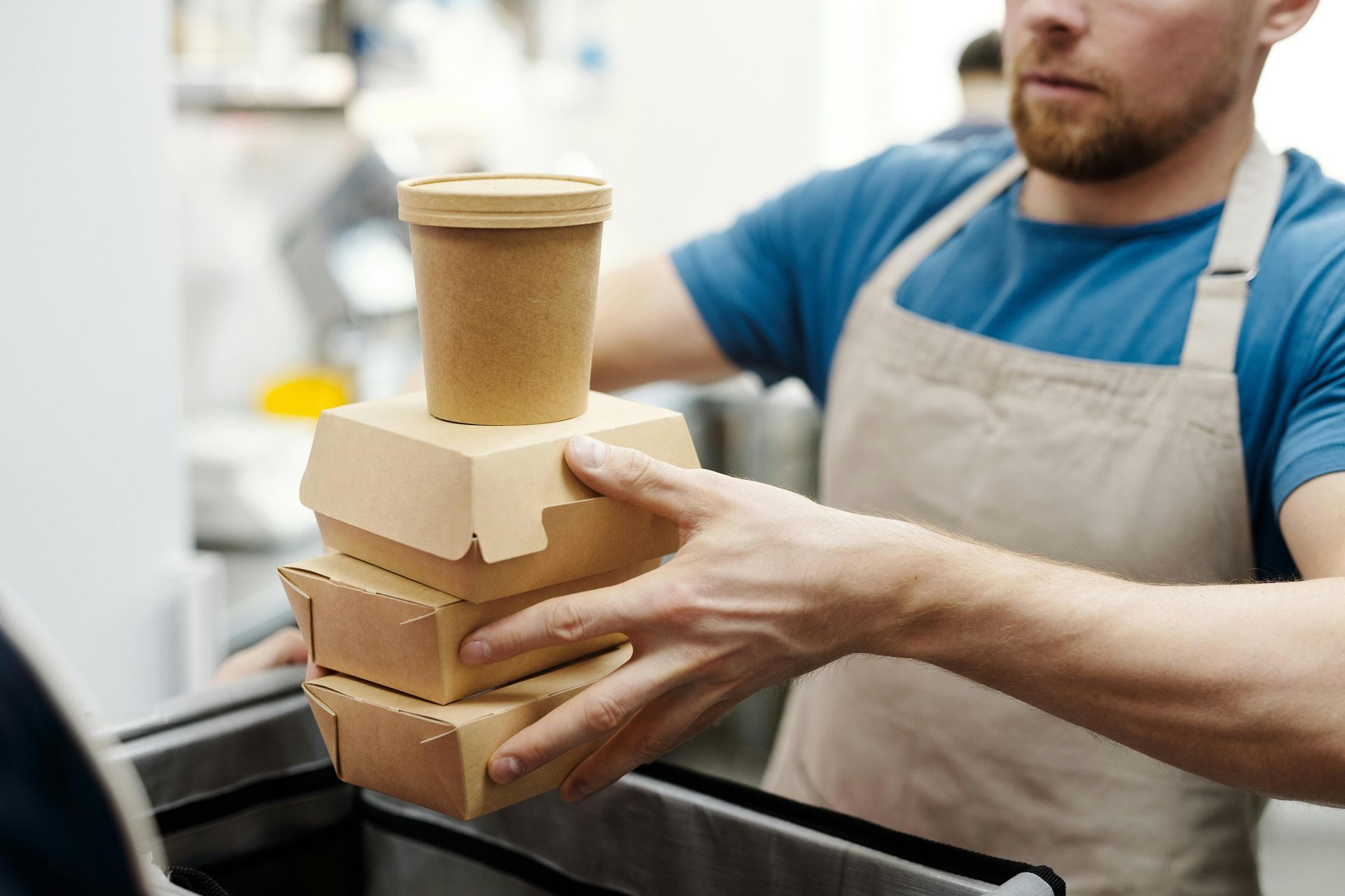 Man in apron holding stacked takeout containers and a coffee cup, likely at a restaurant.