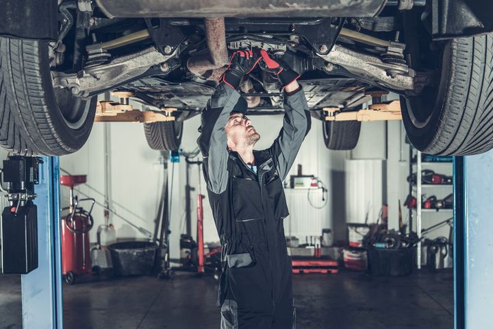 A mechanic in a grey and black uniform stands under a lifted car, performing maintenance in an auto repair shop.