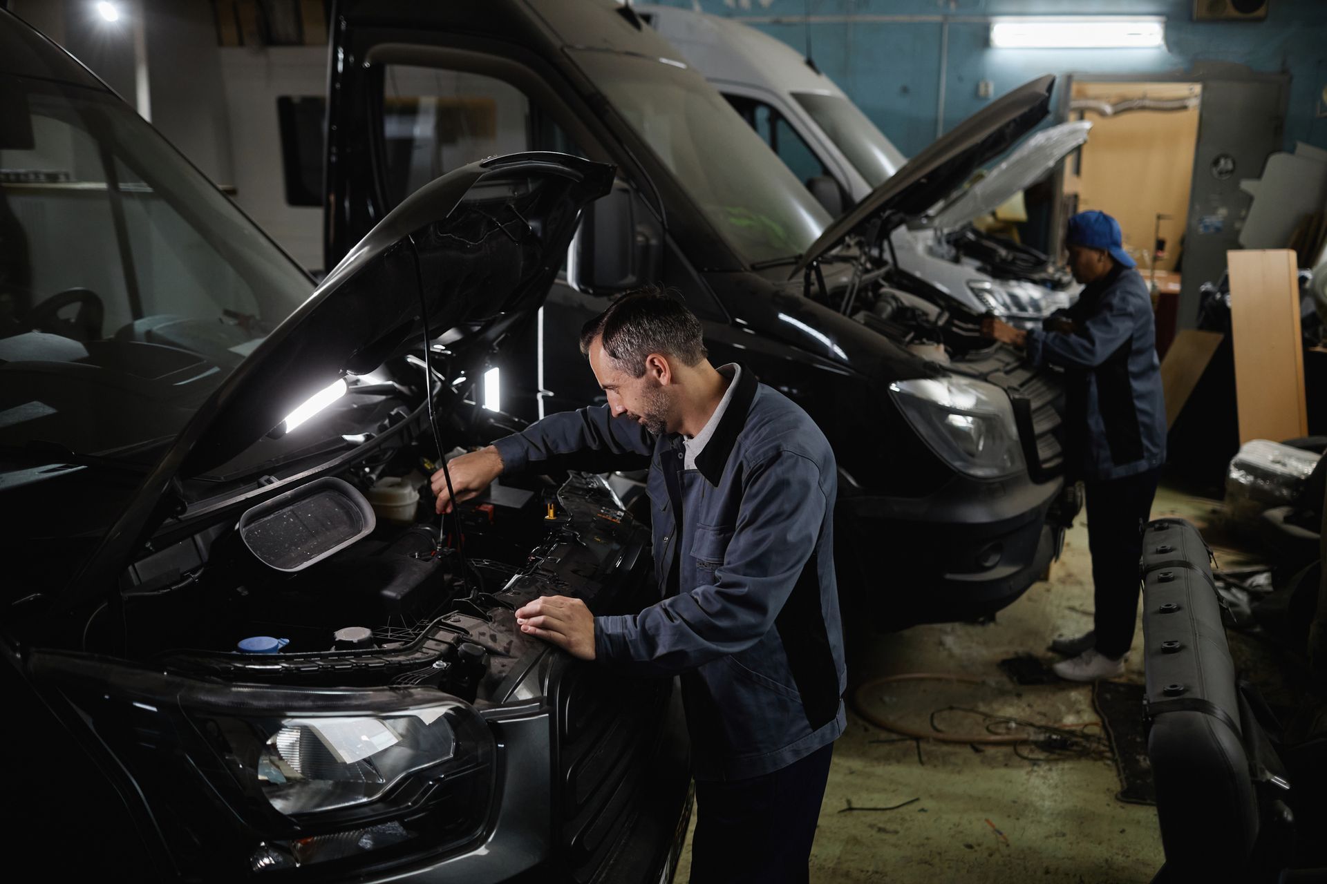 Two workers in blue uniforms repair the engines of two black vans inside a dimly lit workshop.