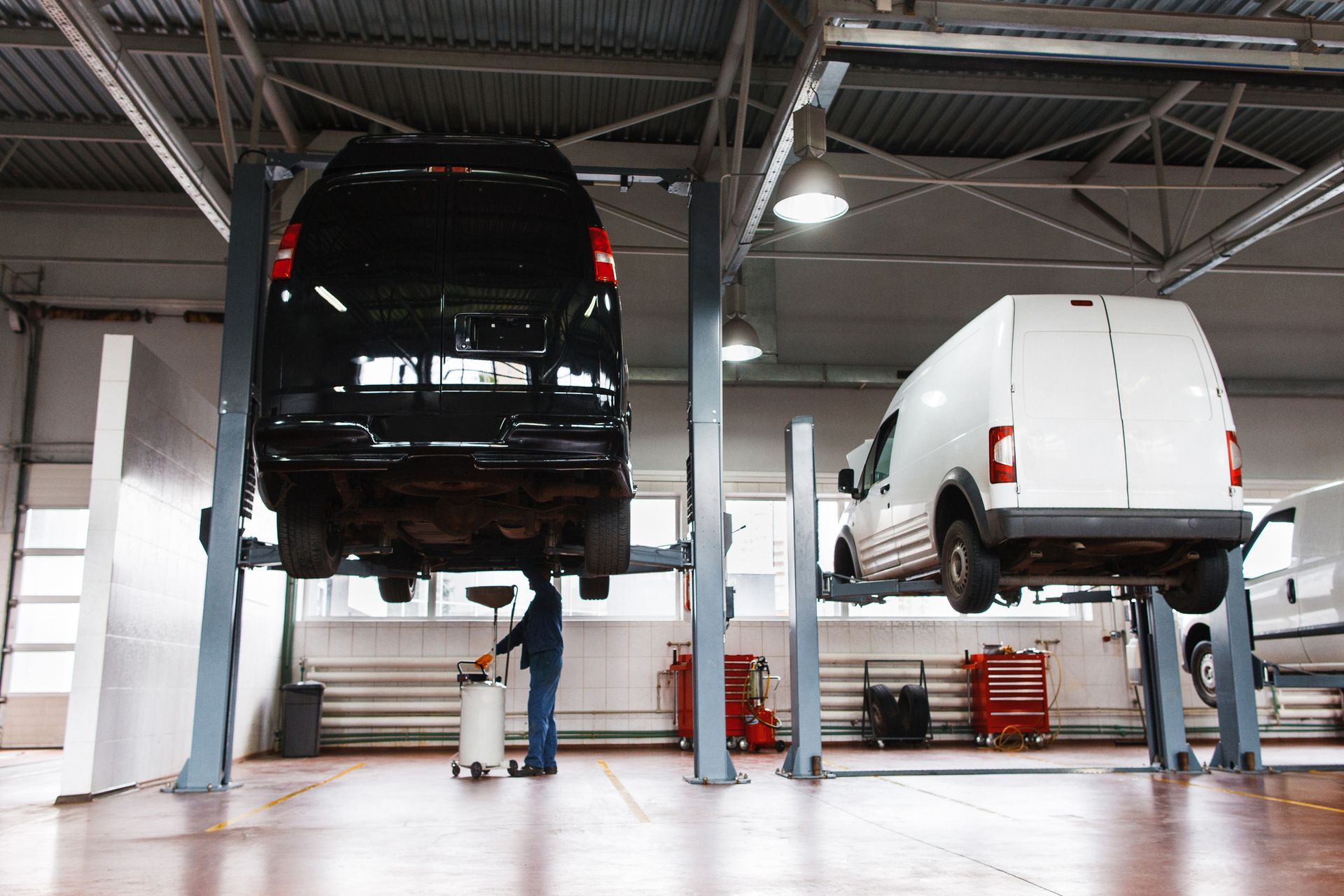 A mechanic works under a lifted black van in a garage, with a white van on a nearby lift.