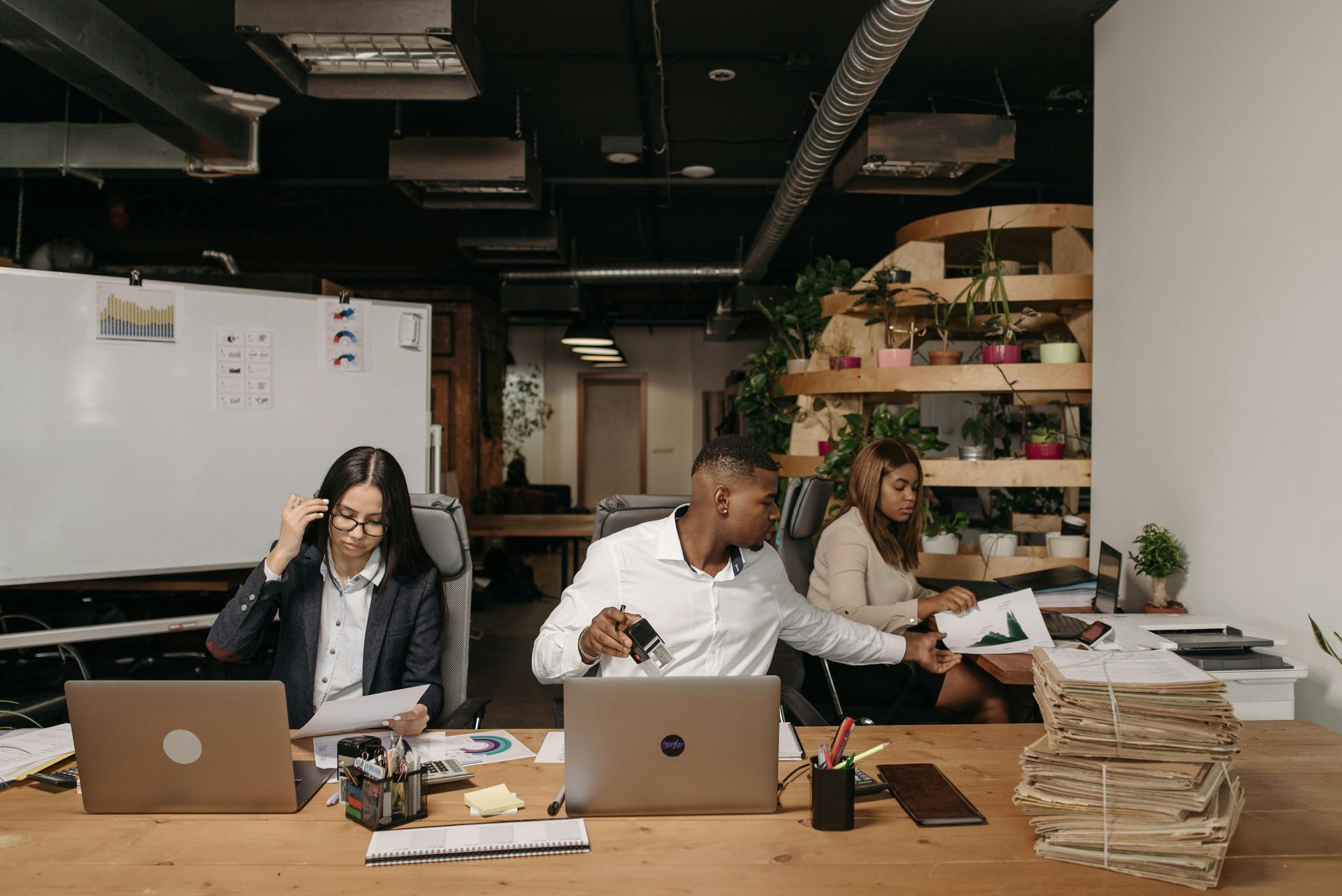 Three office workers sit at a shared wooden desk in an industrial-style workspace, focused on their laptops and documents.