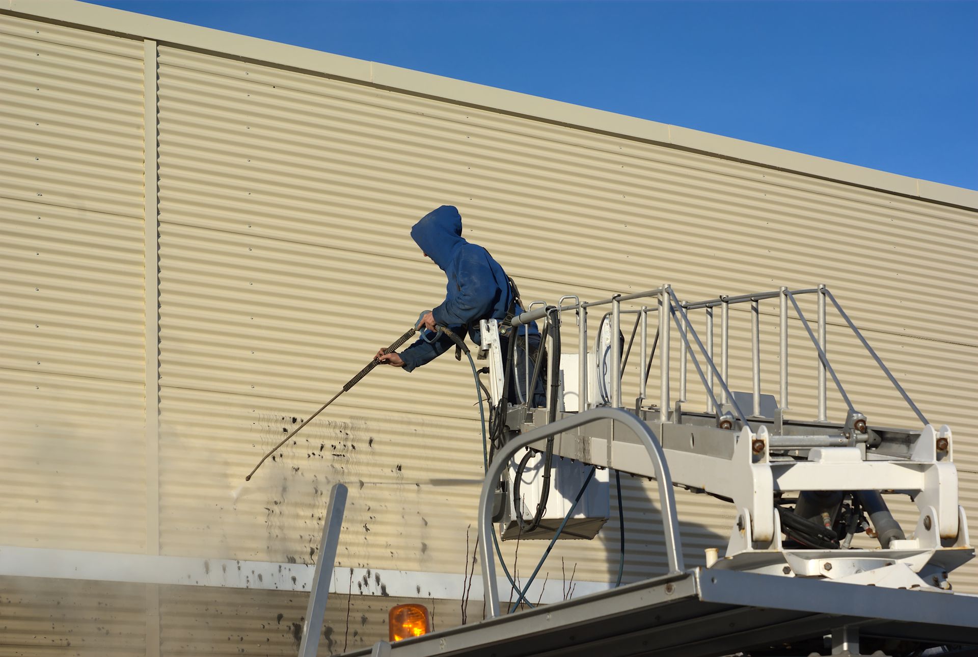Worker using a lift-mounted pressure washer to clean an industrial building exterior.