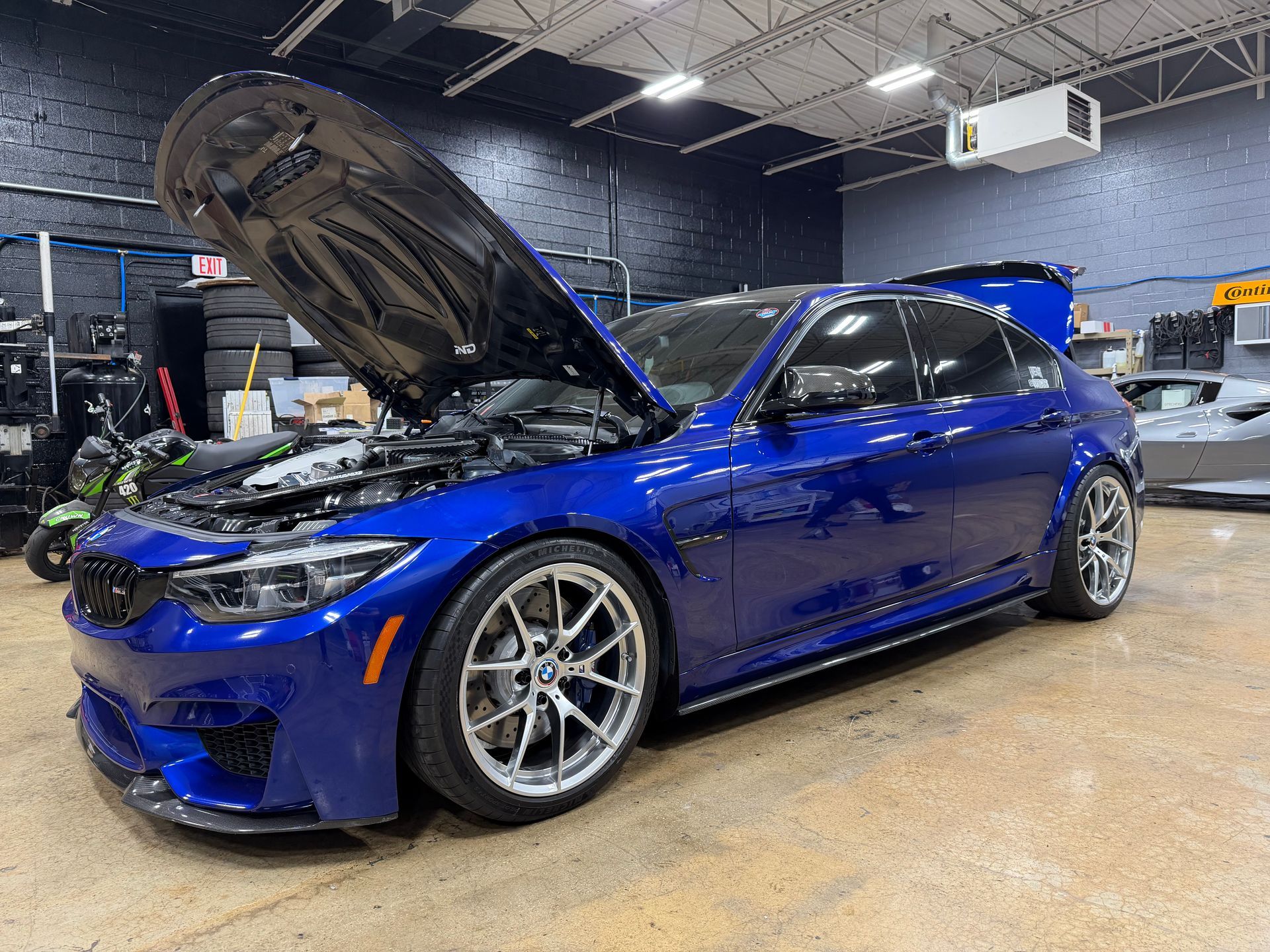 Blue BMW sedan with hood open in a garage, showcasing engine modifications.