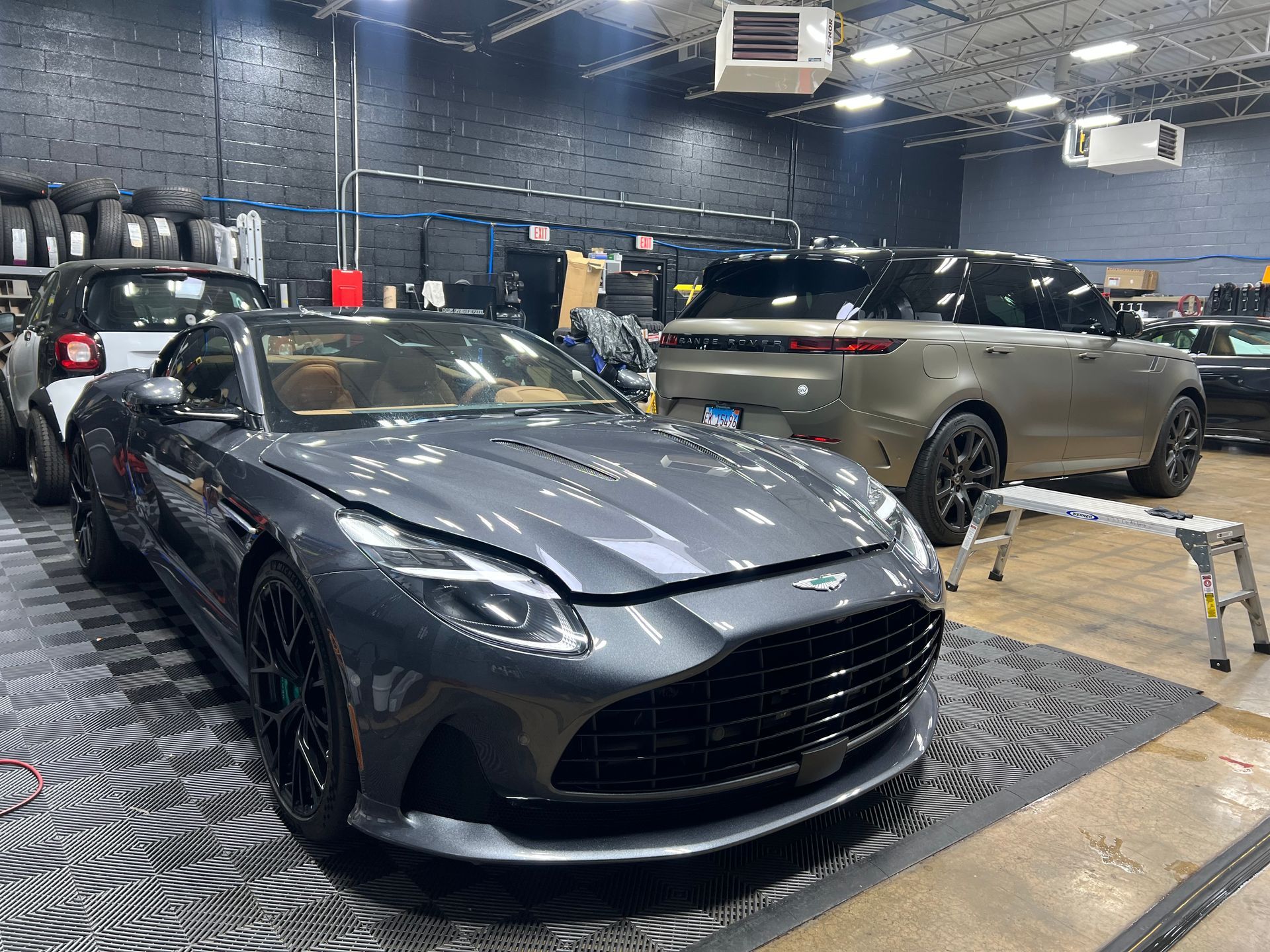 Gray Aston Martin and SUV parked inside a garage, with tires and equipment visible.