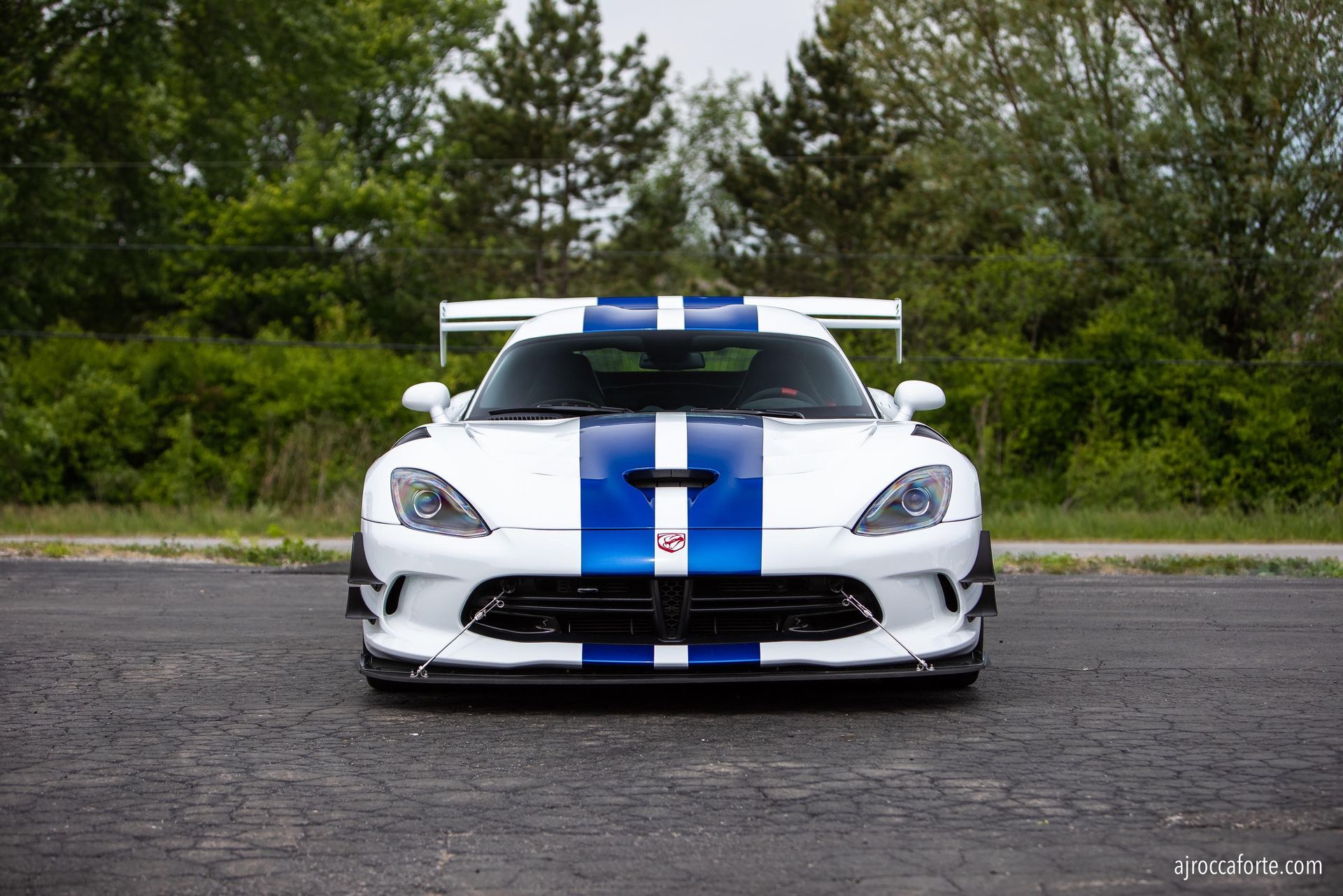 White Dodge Viper with blue racing stripes, front view, parked on asphalt, trees in background.