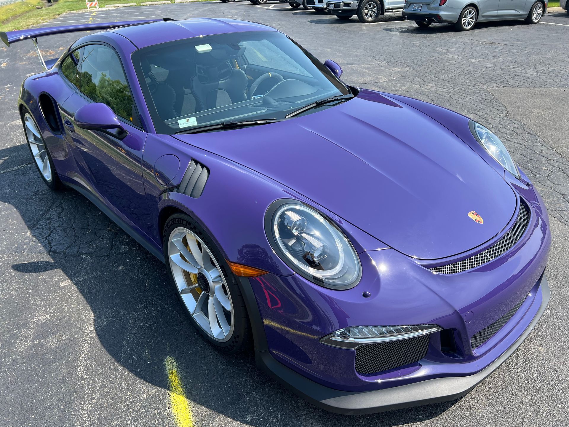 Gray classic Bentley car with white-wall tires parked on asphalt in front of a green treeline.