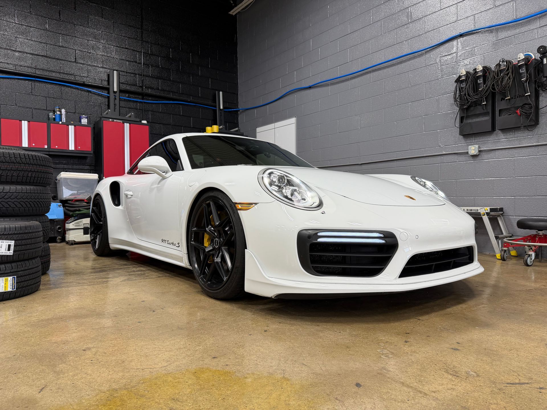 White Porsche sports car inside a garage, black wheels, against a backdrop of tools and equipment.