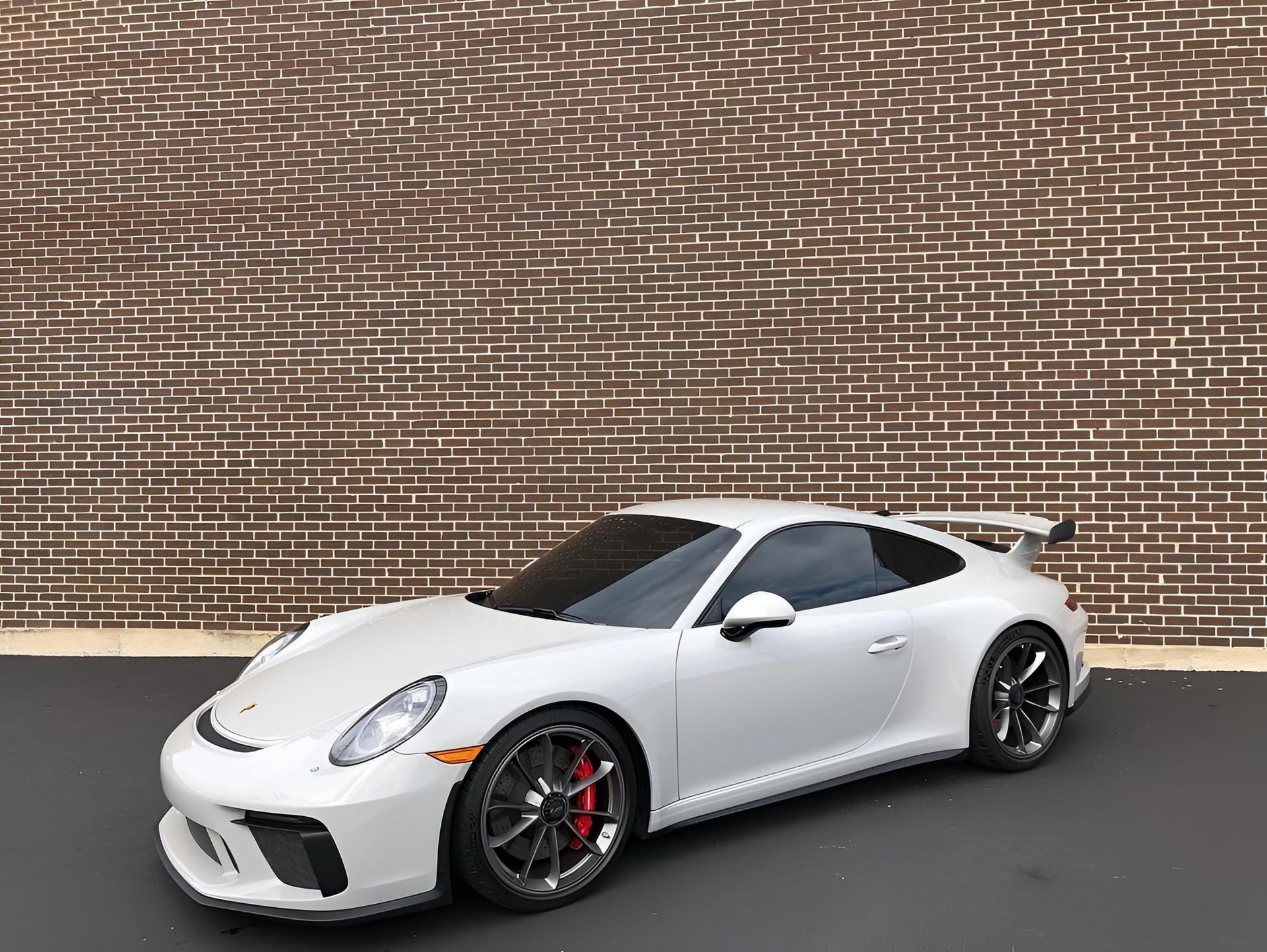White Porsche 911 GT3 parked in front of a brick wall, with tinted windows and black wheels.